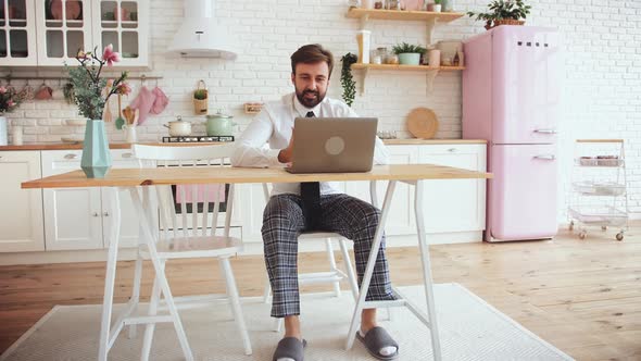 Young Bearded Man Working From Home with Laptop Wearing Shirt Tie and Pajama Pants in Kitchen