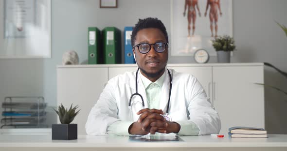 African Male Positive Doctor in Earphones Having Online Consultation with Patient in Clinic Office