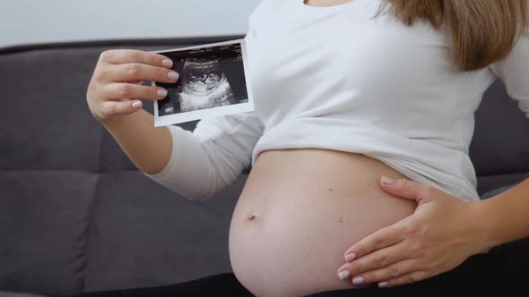 A Pregnant Woman Strokes Her Tummy and Shows a Photo of the Ultrasound Diagnosis of the Fetus