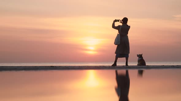 Woman Taking Photo and Standing with Corgi Dog on Sea Beach During Evening Sunset Spbi