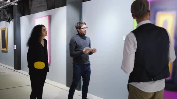 Male Guide Is Talking for Two Visitors of Art Gallery and Showing Pictures