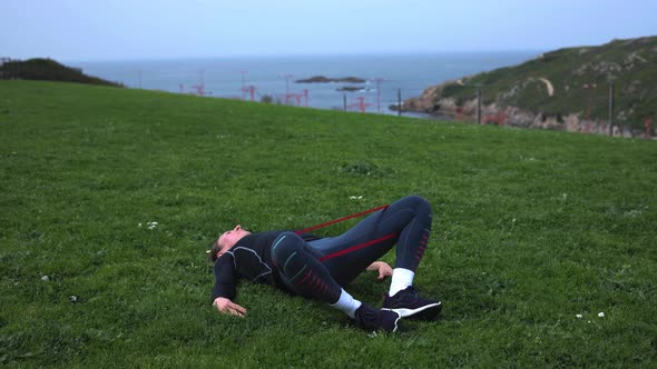 a Girl in Blue Leggings Lying on Green Grass and Doing Exercises with Rubber for the Buttocks