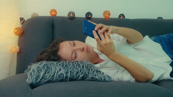 Young Woman is Using Smart Phone and Lying on Couch Leisurely in Living Room