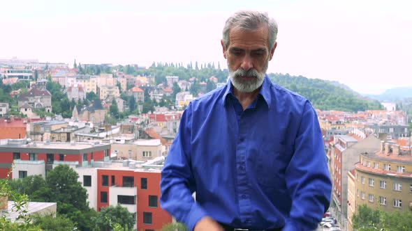 Old Senior Man Points on Watch (Show Time) - City (Buildings) in Background