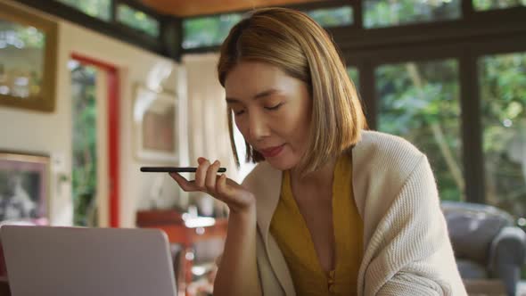 Asian woman sitting at table working from home talking on smartphone and using laptop