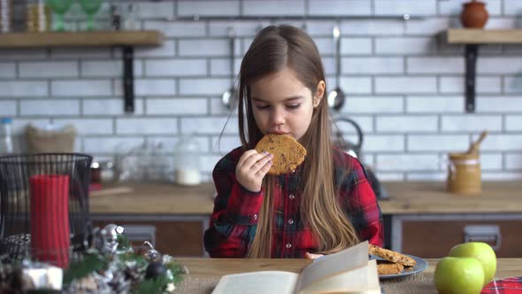 Small Beautiful Girl Sit on the Kitchen Table, Eating Cookies and Leafing Through Book