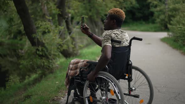Curios African American Man in Wheelchair Taking Photo of Nature on Smartphone in Summer Forest