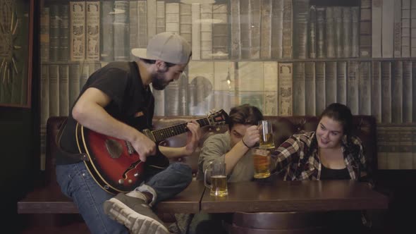 Young Positive Bearded Man Playing Guitar in the Bar, His Friends Sitting Near Drinking Beer