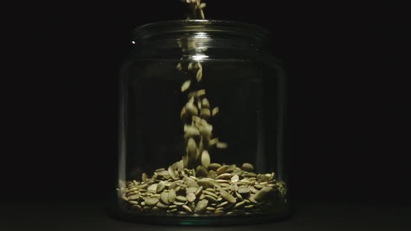 Peeled Pumpkin Seeds Fall Into Empty Glass Jar On Black Background - Front View