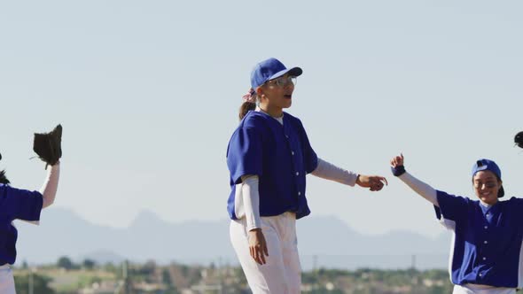 Happy diverse team of female baseball players celebrating after game, embracing and throwing gloves