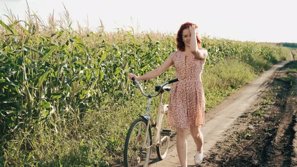 Girl in Stylish Outfit is Walking with Vintage Retro Bike on Countryside Road in Sunshine