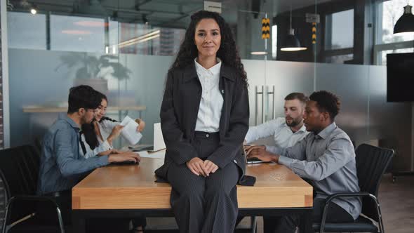 Satisfied Smiling Young Hispanic Businesswoman Sitting Carelessly on Table in Boardroom Multiracial