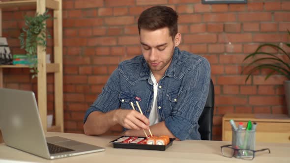 Handsome Male Freelancer Eating Sushi Rolls While Reading News on Laptop in Desk at Home Office