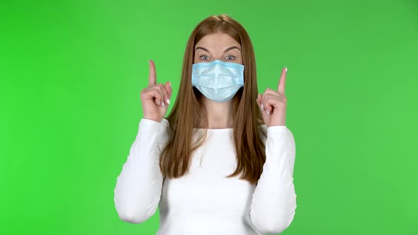 Portrait of Pretty Younggirl in Medical Protective Face Mask Looking at Camera and Pointing Up