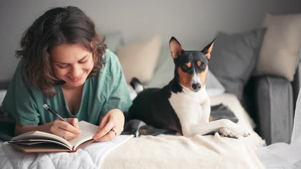 A Young Woman Is Writing a Diary While Lying in Bed with Her Basenji Dog