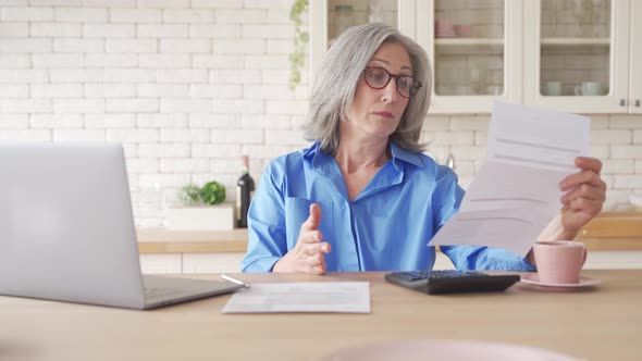 Serious Worried Mature Woman Reading Paper Bill To Pay Online at Home