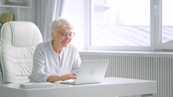 Smiling aged lady with grey hair and glasses types on laptop