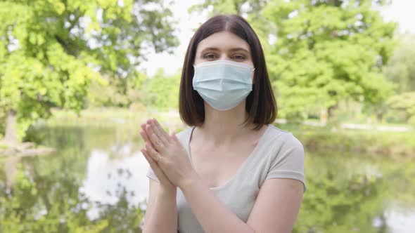 A Young Caucasian Woman in a Face Mask Applauds to the Camera in a Park