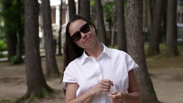 Portrait of a Brunette in a White Dress Against the Background of Tree Trunks and a House with