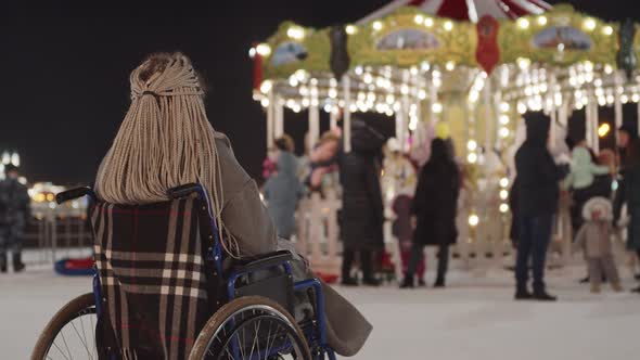 A Young Sad Woman in a Wheelchair Looks at a Bright Carousel Outdoors