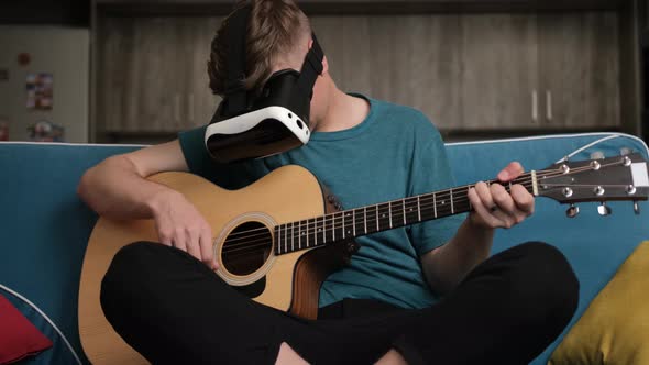 Young Man Sitting on Sofa with the Virtual Reality Glasses Is Playing at the Guitar