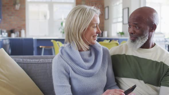 Happy senior diverse couple in living room sitting on sofa, using smartphone, making video call