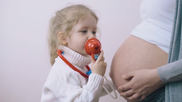 Girl with Toy Stethoscope Kisses Pregnant Mom Tummy in Room