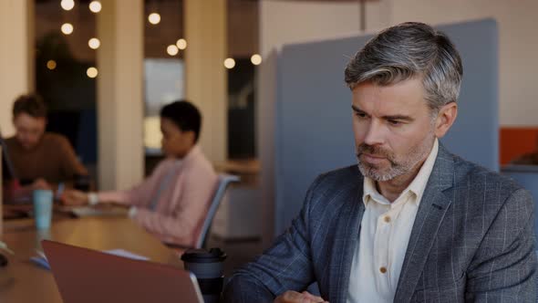 Caucasian Senior Serious Man Working on the Laptop While Sitting at the Table on the Office Center