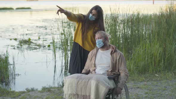 Portrait of Confident Young Woman in Face Mask Talking with Paralyzed Man in Wheelchair. Wide Shot