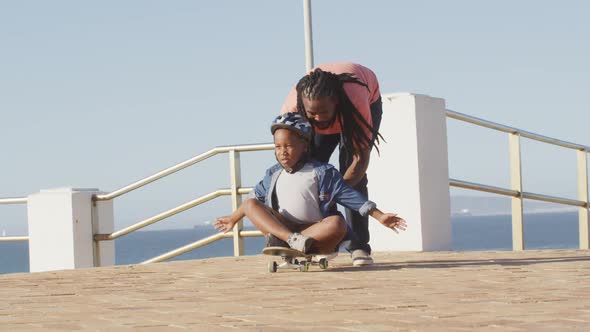Video of happy african american father learning son how to skateboard on promenade