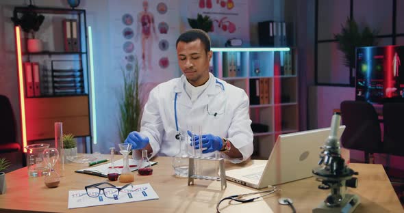 Male Chemist in Uniform Working on Laptop and Viewing Chemical Solutions in Flasks During His Work