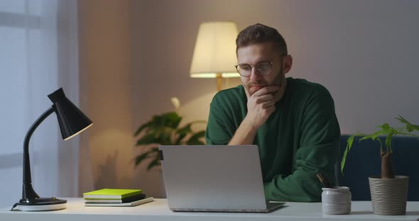 Thoughtful Young Man Is Sitting at Table with Laptop Writer or Journalist Is Working on Book in Home