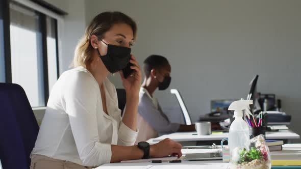 Two diverse female colleagues wearing face mask, working at desk in office