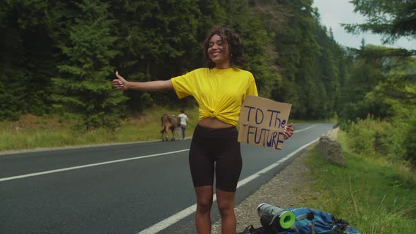 Pretty Smiling African American Woman Tourist with Cardboard and Thumbs Up Hitchhiking at Road