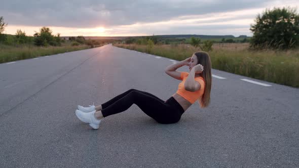 Athletic Girl in Sportswear Doing Abs Exercises Flexing Legs on Road at Nature