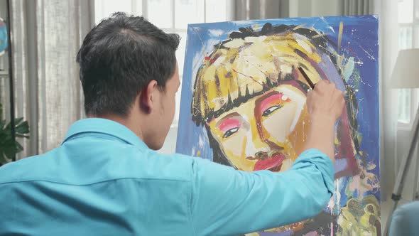 Hind View Of An Asian Artist Man Holding Paintbrush Painting A Girl's Hair On The Canvas