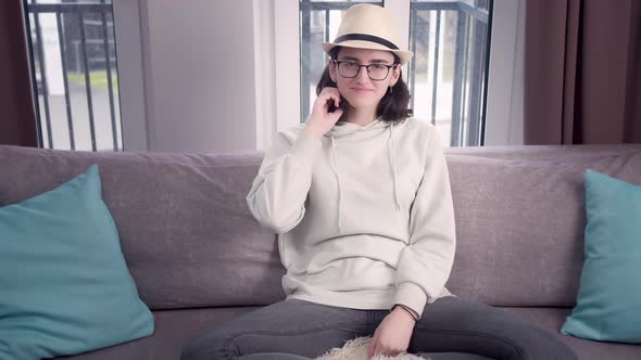 Pretty girl in glasses and hat looks straight at the camera sitting cross-legged on the couch