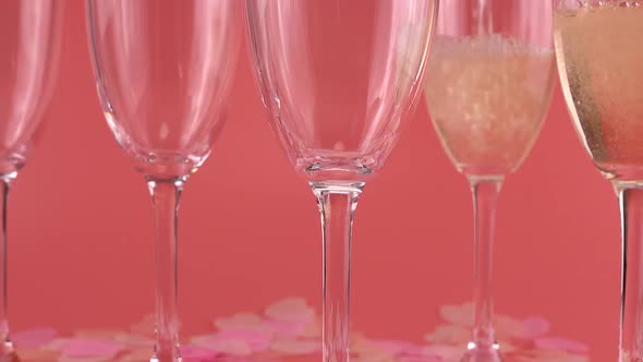 Pouring sparkling wine into glasses on a pink background with heart-shaped confetti.