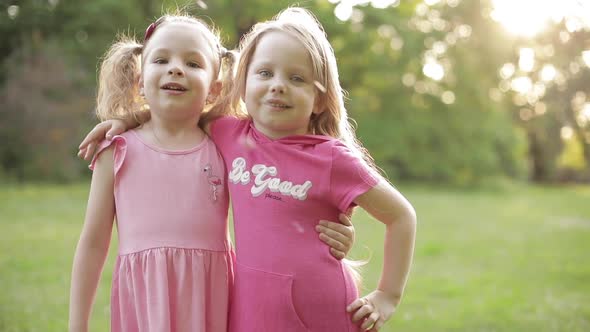 Cute Young Children in Pink Dresses Hugging and Smiling at Camera in Park