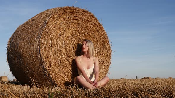 blonde girl sits by a rolled haystack in field in sunset time