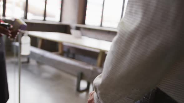 Midsection of woman at bar, holding thermometer, using sanitising spray on hands of male colleague