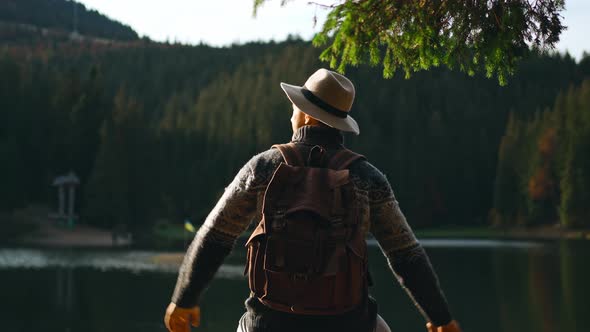 Rear View Traveler Man Wearing Hat with Backpack Standing By Mountain Lake with Calm Blue Water