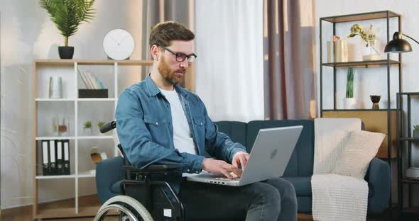 Man in Glasses Sitting in Wheelchair Working on Laptop at Home and Looking at Camera