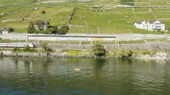 Aerial orbit around kayak on Lake Leman with swiss train passing along the shore. Lavaux vineyard -
