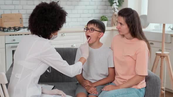 Afro American Woman Doctor in Mask Looks at Sick Child's Throat While Examining at Home Next to Mom