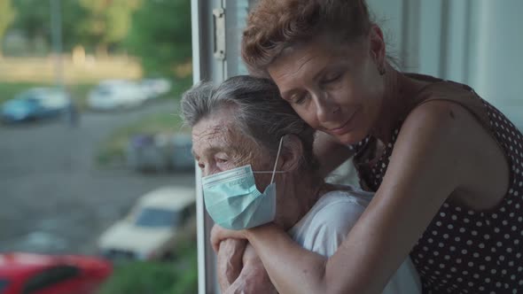 Adult Daughter Embracing with Loving Senior Mother 90 Years in Mask Standing at Window Relatives