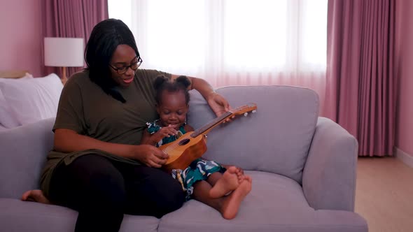 Close up African American mother play ukulele with her child on sofa in bedroom