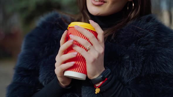 Cup of hot coffee in woman's hands outdoors. Young girl in fur coat warming her hands on the plastic