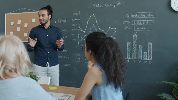 Joyful Arab Man Speaking to Colleagues Pointing at Chalkboard Making Report in Workplace