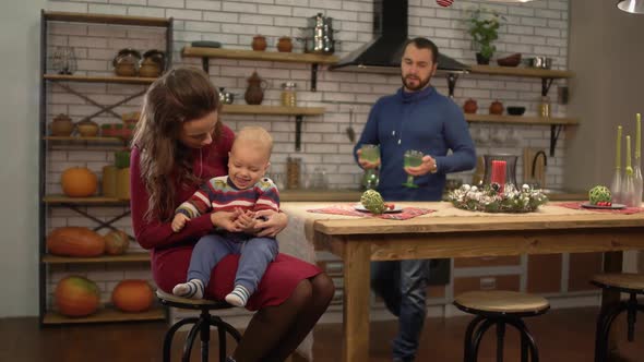 Young Pretty Woman Sitting at the Table in the Kitchen with Baby on Her Pals While Handsome Bearded